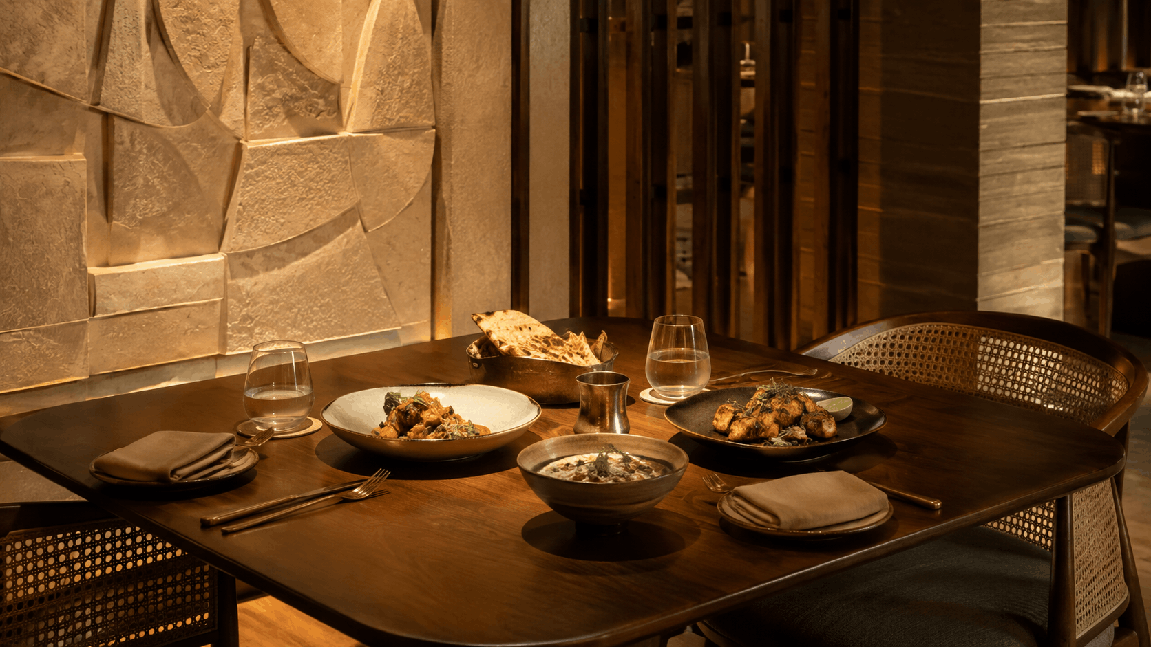 Close-up of a warmly lit dining table with plated food, water glasses, and naan bread against a textured stone wall